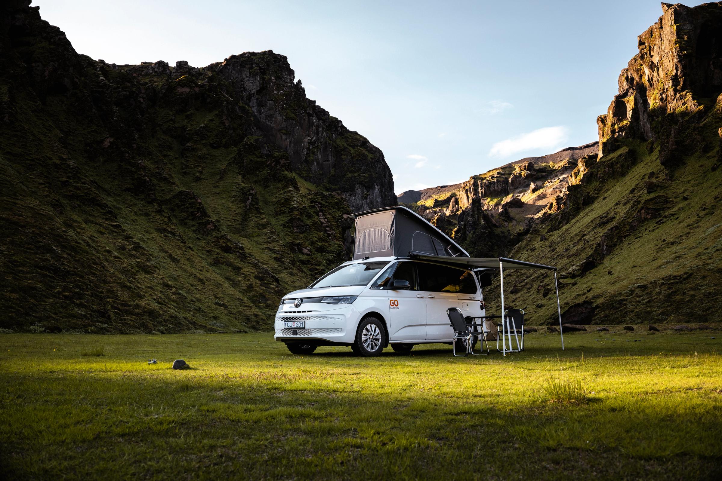 A white camper van with a raised pop-top roof and extended awning, set up with a table and chairs, sits in a lush green mountain valley.