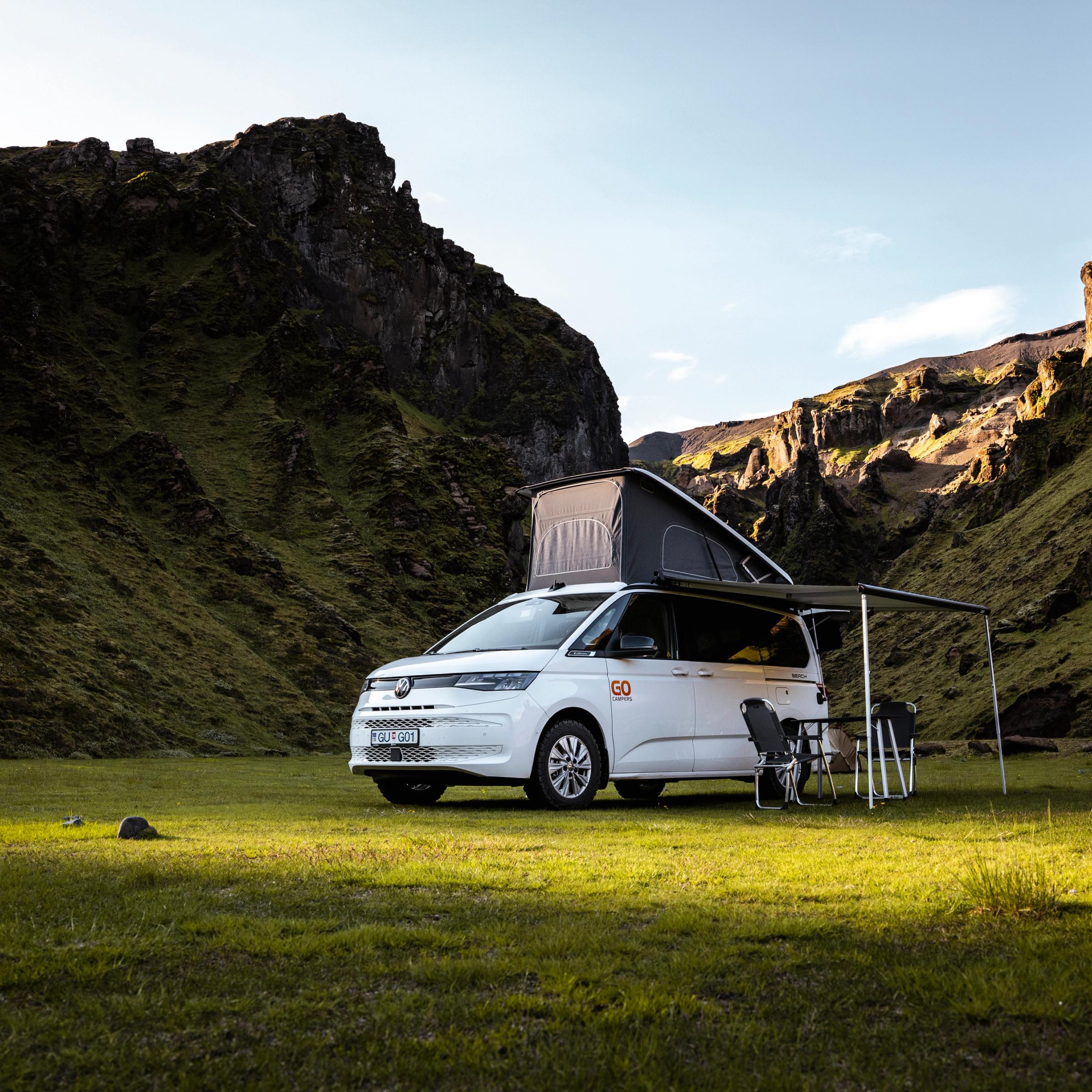 A white camper van with a raised pop-top roof and extended awning, set up with a table and chairs, sits in a lush green mountain valley.