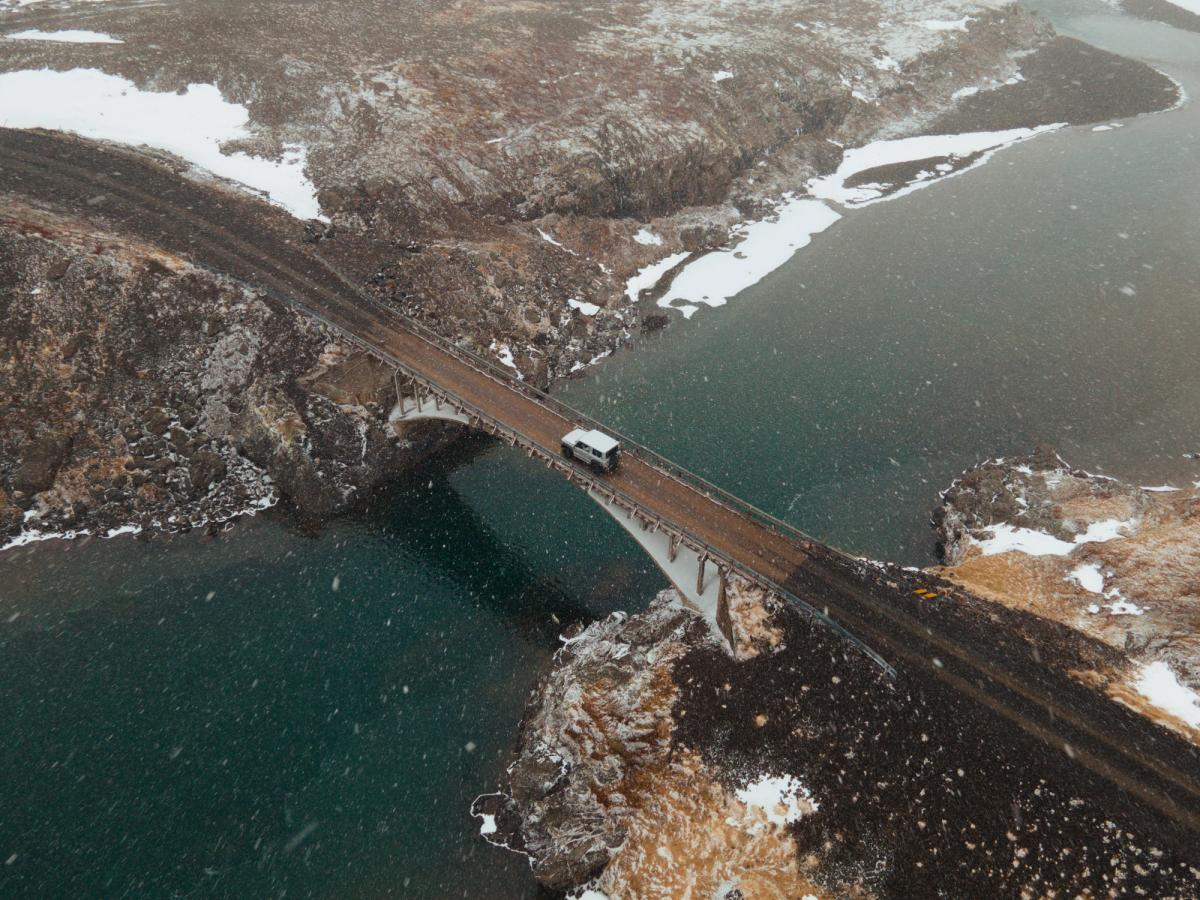 Aerial view of a white van driving across a bridge over water in a snowy, rocky landscape.