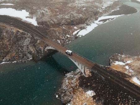 Vista aérea de una furgoneta blanca cruzando un puente sobre el agua en un paisaje nevado y escarpado, con nieve cayendo.