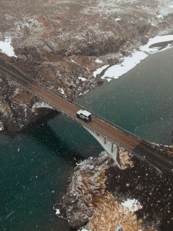 Aerial view of a white van crossing a bridge over water in a snowy, rugged landscape with falling snow.