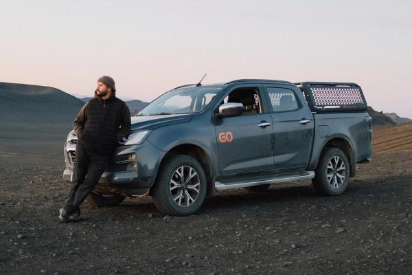 A man leans on a dark pickup truck in a desolate, rocky landscape at dusk.