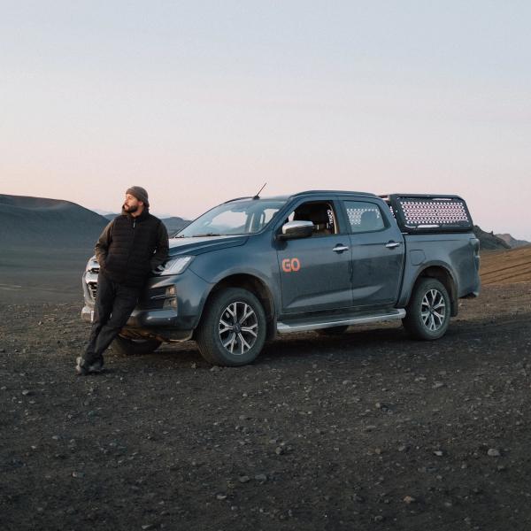 A man leans against a dark grey pickup truck in a barren, rocky landscape under a pale sky.