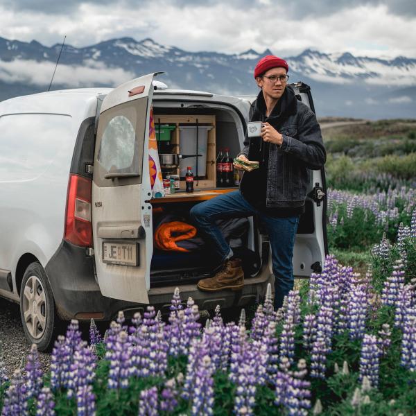 a man standing in a field of purple flowers next to a white van with a license plate that says f.e.ftn