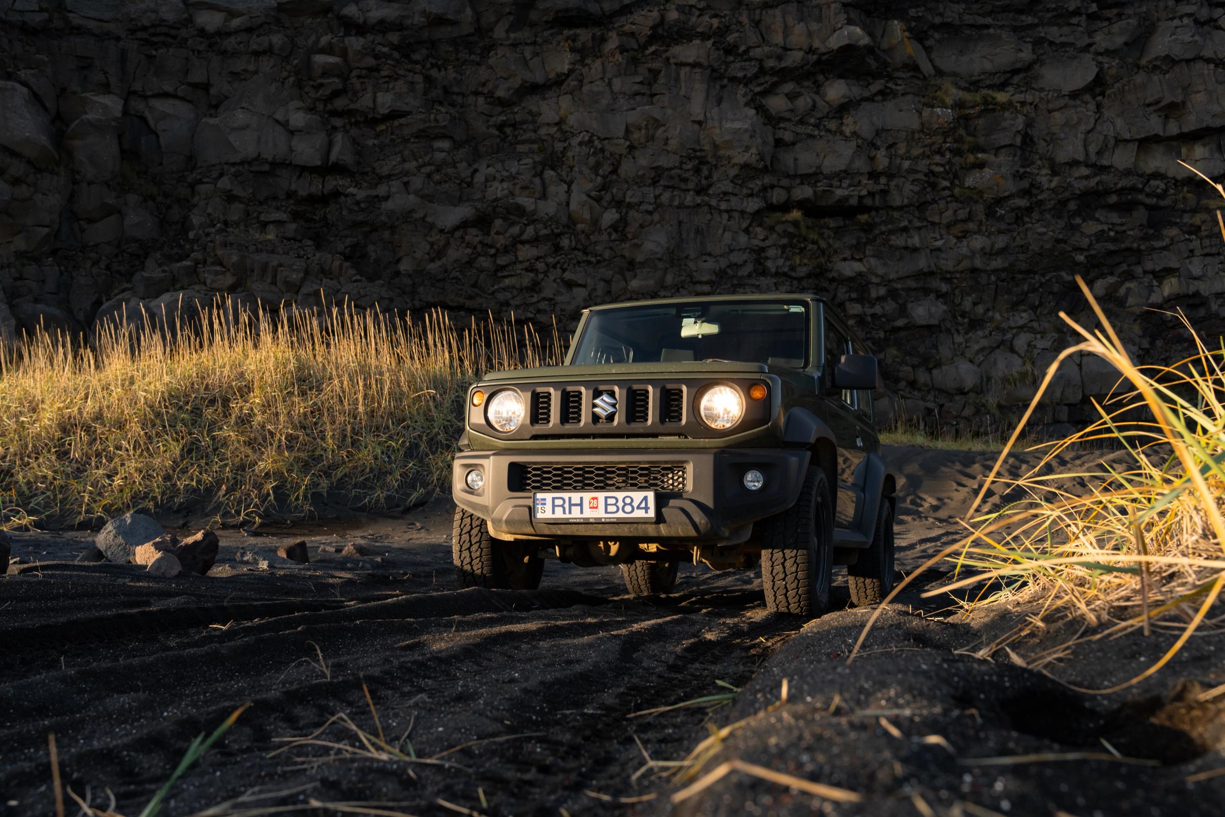 Suzuki Jimny mit Schaltgetriebe in der Frontansicht, mit eingeschalteten Scheinwerfern auf dunklem Untergrund unterhalb einer Felswand