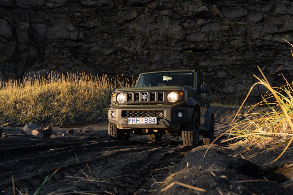 Suzuki Jimny mit Schaltgetriebe in der Frontansicht, mit eingeschalteten Scheinwerfern auf dunklem Untergrund unterhalb einer Felswand