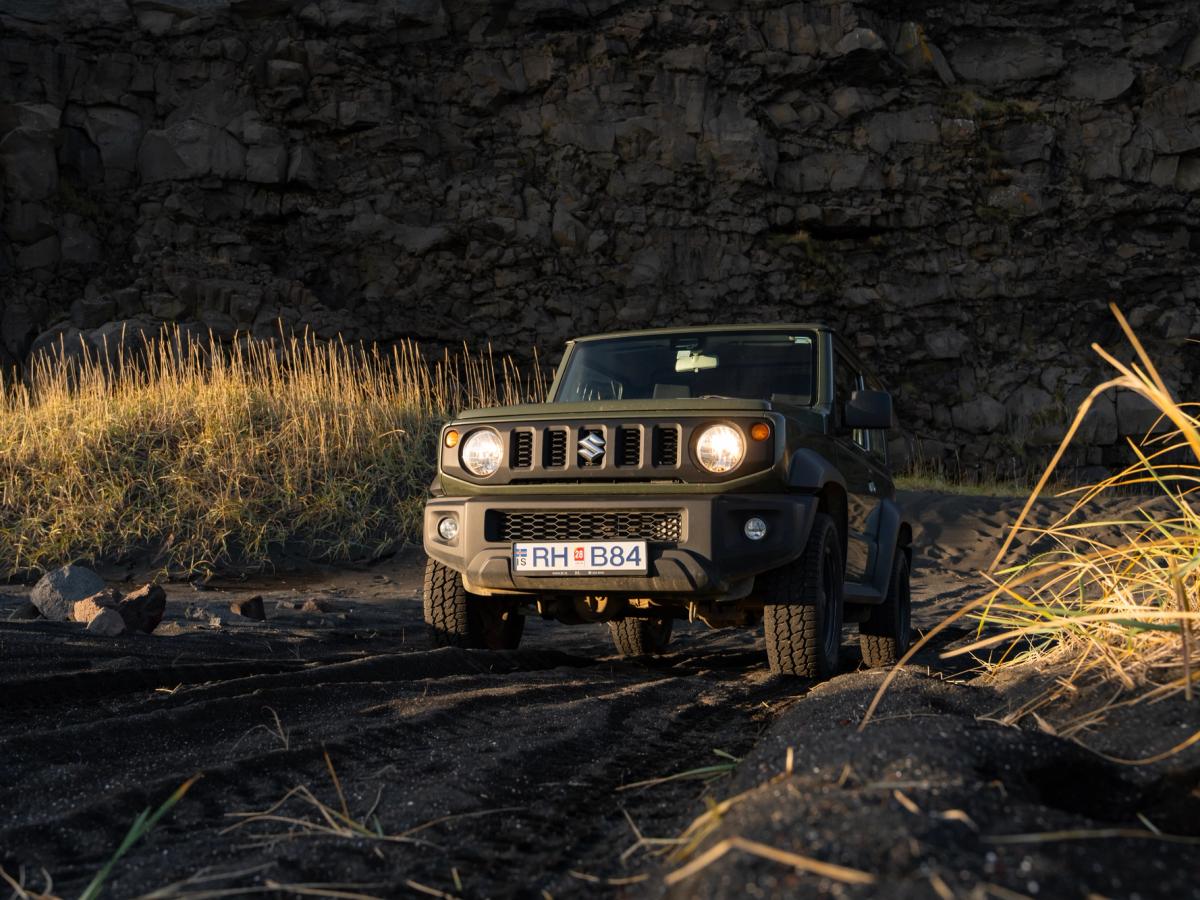 Jimny Front Dark Terrain Suzuki Jimny mit Schaltgetriebe in der Frontansicht, mit eingeschalteten Scheinwerfern auf dunklem Untergrund unterhalb einer Felswand