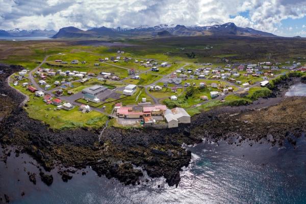 Aerial view of a small village on a rocky peninsula, surrounded by blue sea, green land, and distant mountains.