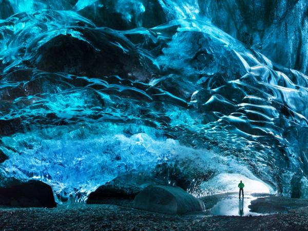 A person stands within a majestic blue ice cave, its walls reflecting the light.