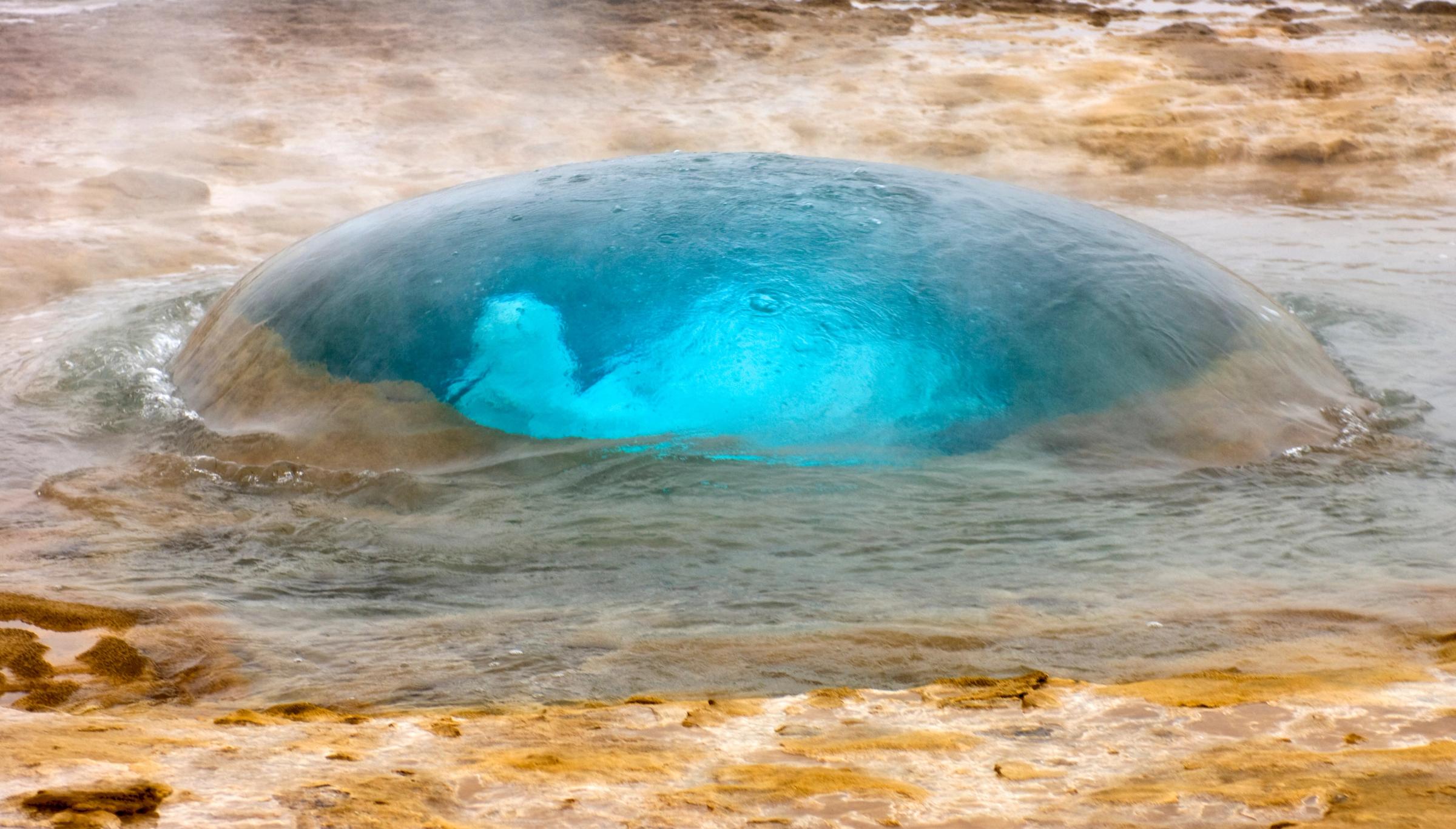 A large, glowing blue bubble of water forms in a geyser pool, surrounded by steam.