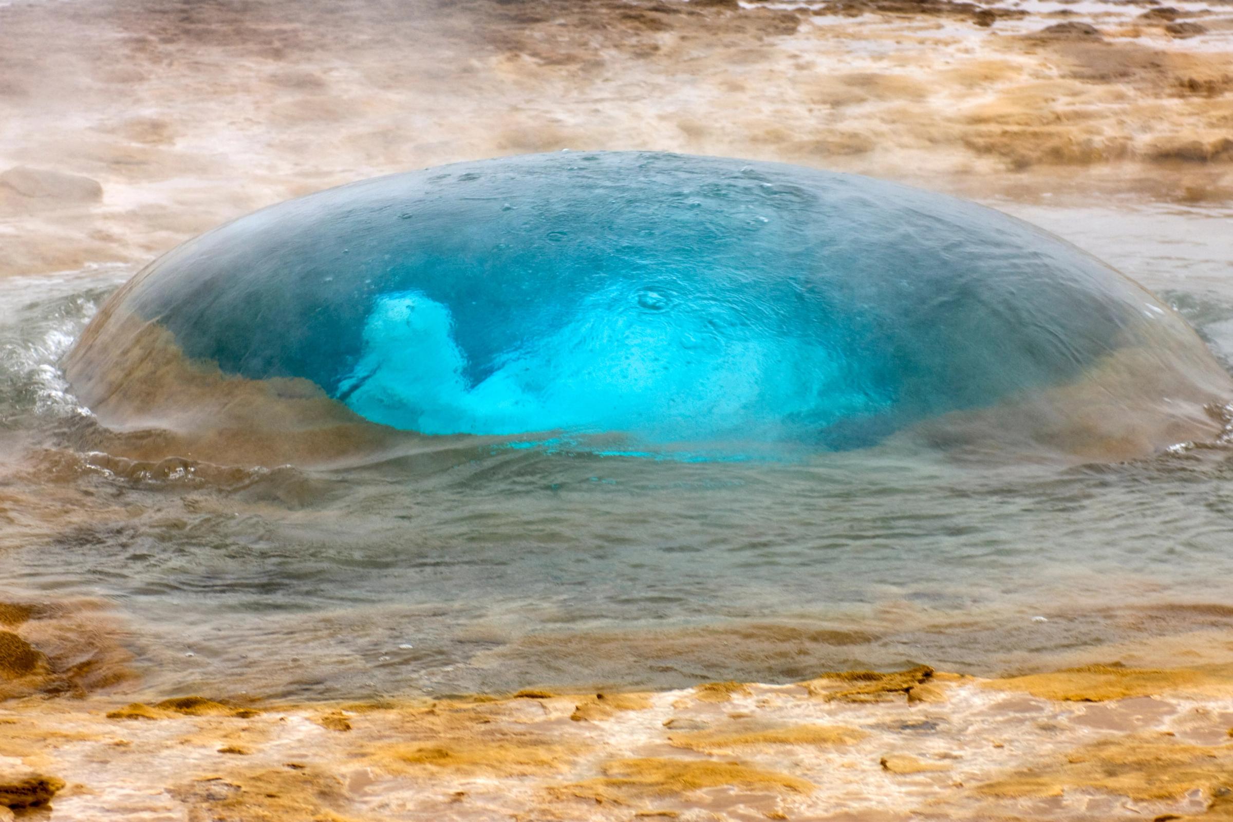 Una gran burbuja de agua azul brillante se forma en una poza de géiser, rodeada de vapor.