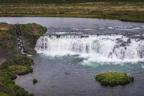 A wide waterfall cascades into a river, with a tiered fish ladder on the left bank and a small grassy island.