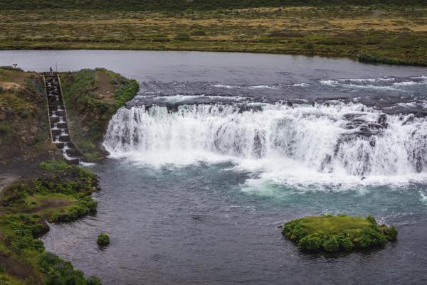 A wide waterfall cascades into a river, with a tiered fish ladder on the left bank and a small grassy island.