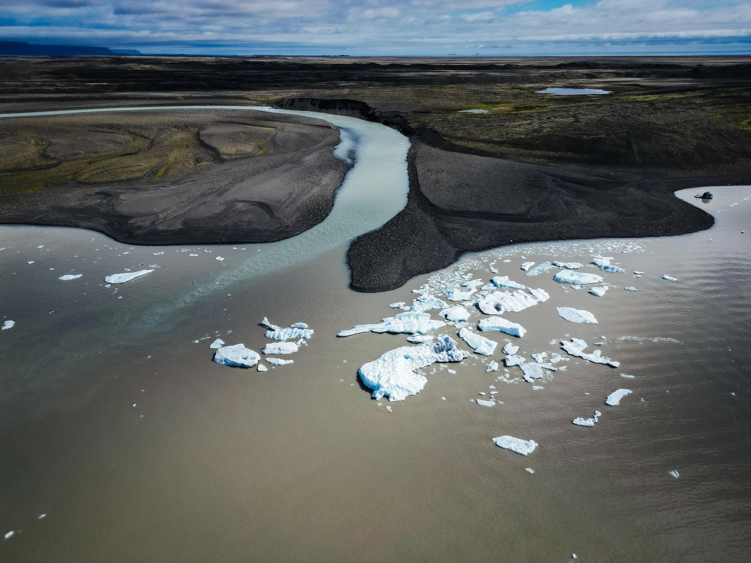 Aerial view of a glacial river flowing into a lake with icebergs, surrounded by dark, barren land.