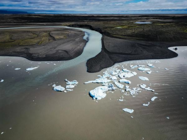 Aerial view of a glacial river flowing into a lake with icebergs, surrounded by dark, barren land.