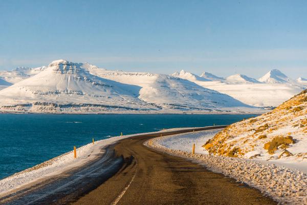 Carretera serpenteante junto a un fiordo con montañas cubiertas de nieve bajo un cielo despejado.