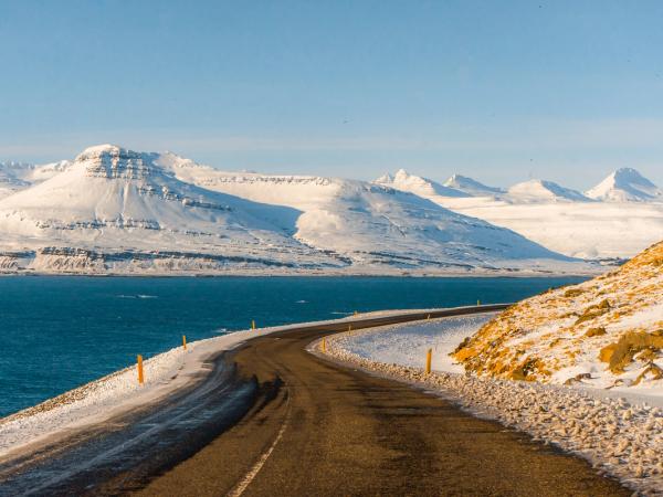Una carretera sinuosa junto a un fiordo azul con montañas nevadas en la distancia.
