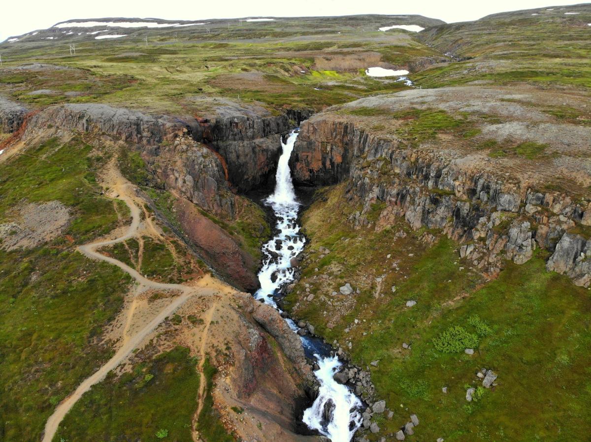 Aerial view of a waterfall plunging into a rocky gorge surrounded by green hills, with a winding dirt path.