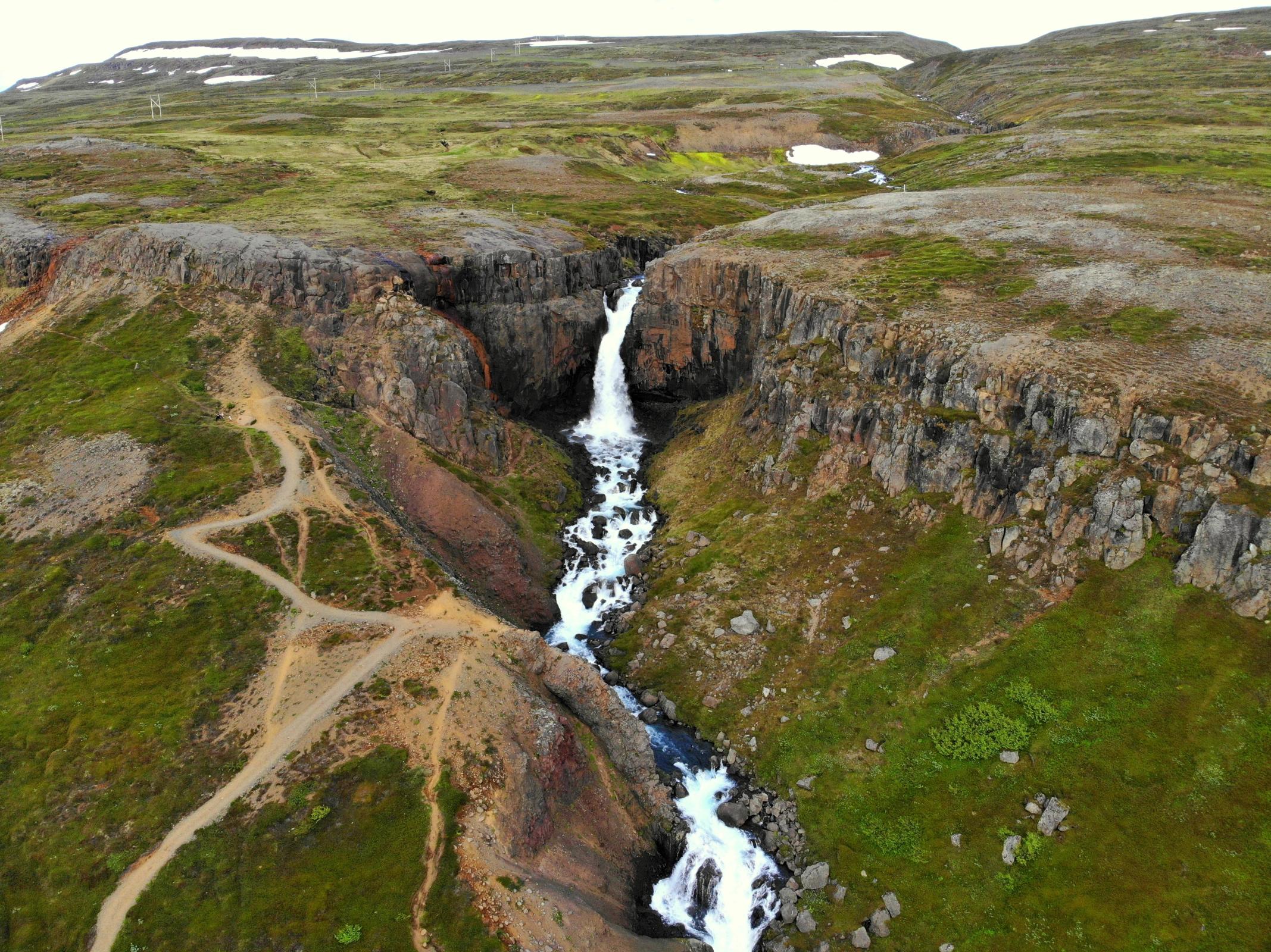 Aerial view of a waterfall cascading through a rocky gorge with a winding dirt path on a green slope.