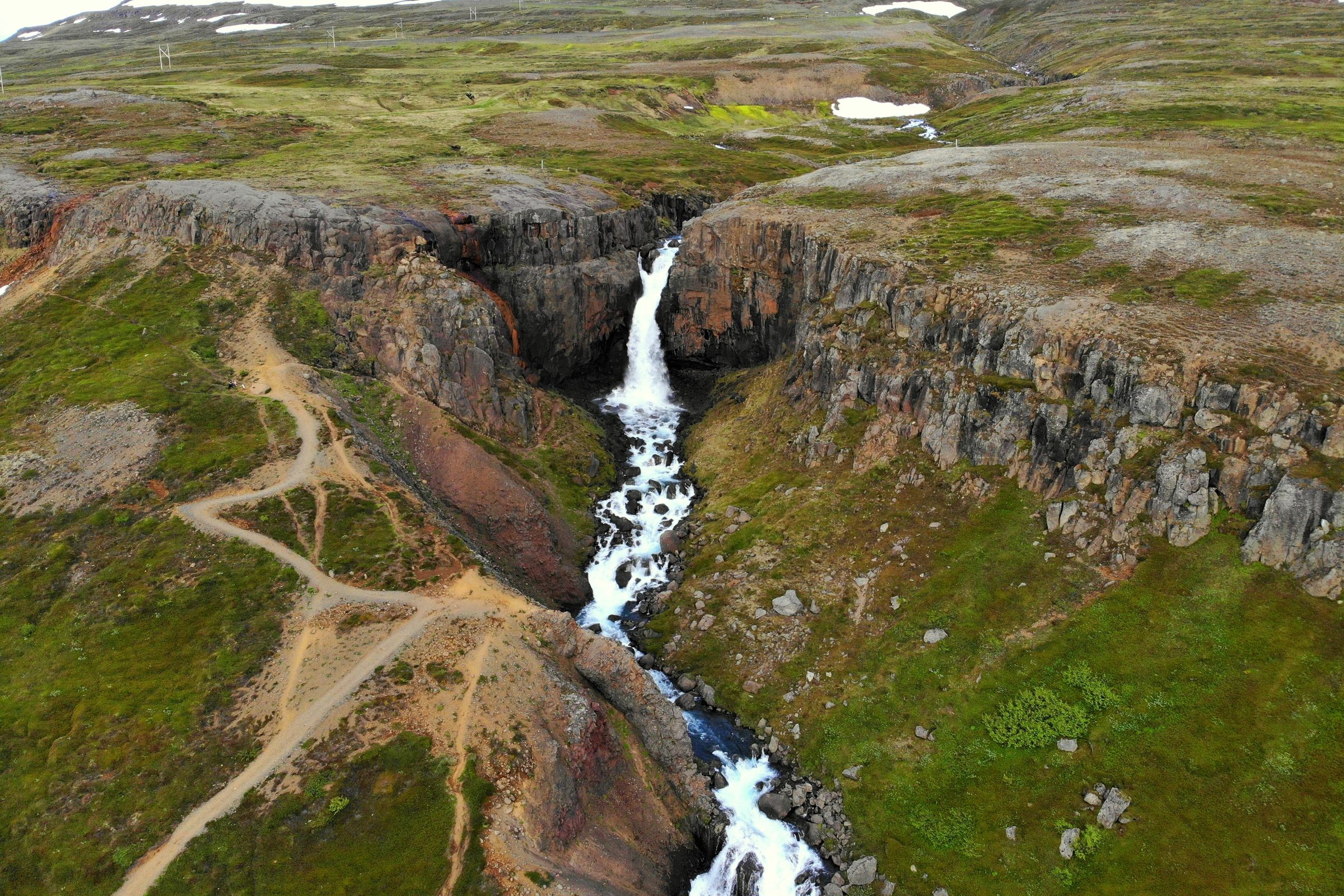 Aerial view of a waterfall cascading through a rocky gorge with a winding dirt path on a green slope.