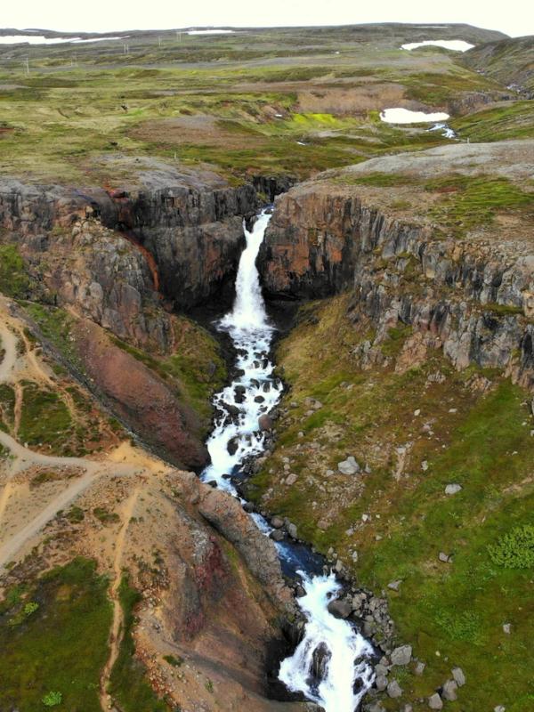 Aerial view of a waterfall plunging into a rocky gorge surrounded by green hills, with a winding dirt path.