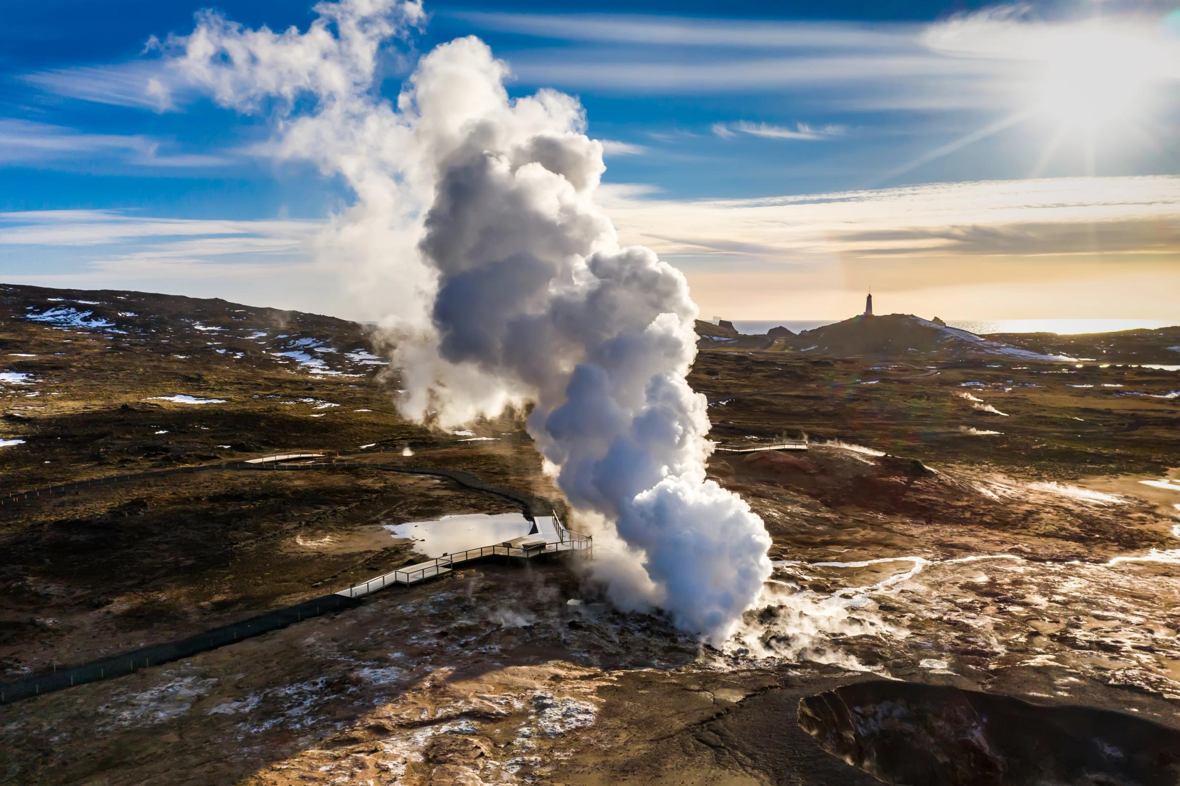 Steam erupts from a geyser in a rocky landscape with a distant monument under a sunny sky.