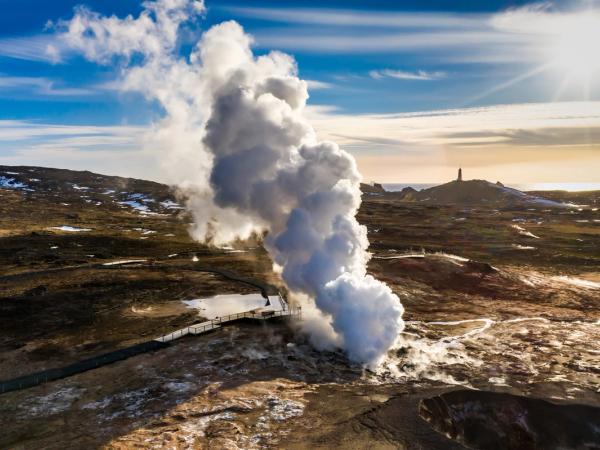 Steam erupts from a geyser in a rocky landscape with a distant monument under a sunny sky.