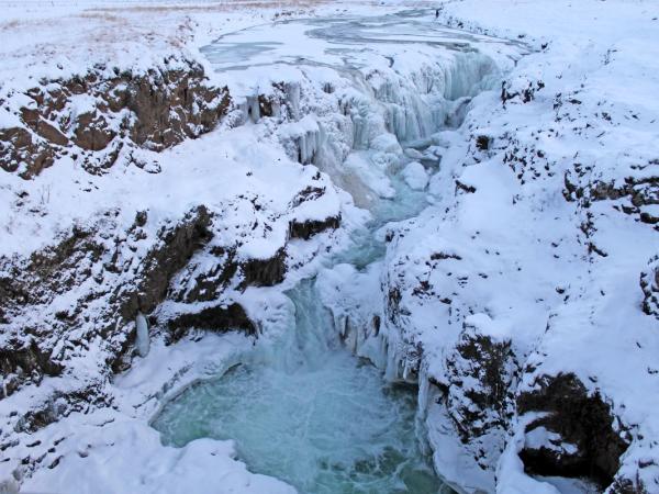 A light blue river flows through a snow-covered canyon, with ice clinging to the rocky cliffs.