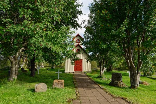 A small, light-colored church with a red door and steeple stands at the end of a paved path, flanked by green grass, trees with red berries, and white gravestones.