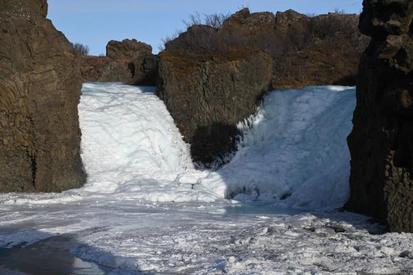 Two partially frozen waterfalls cascade between dark rock cliffs into an icy river.