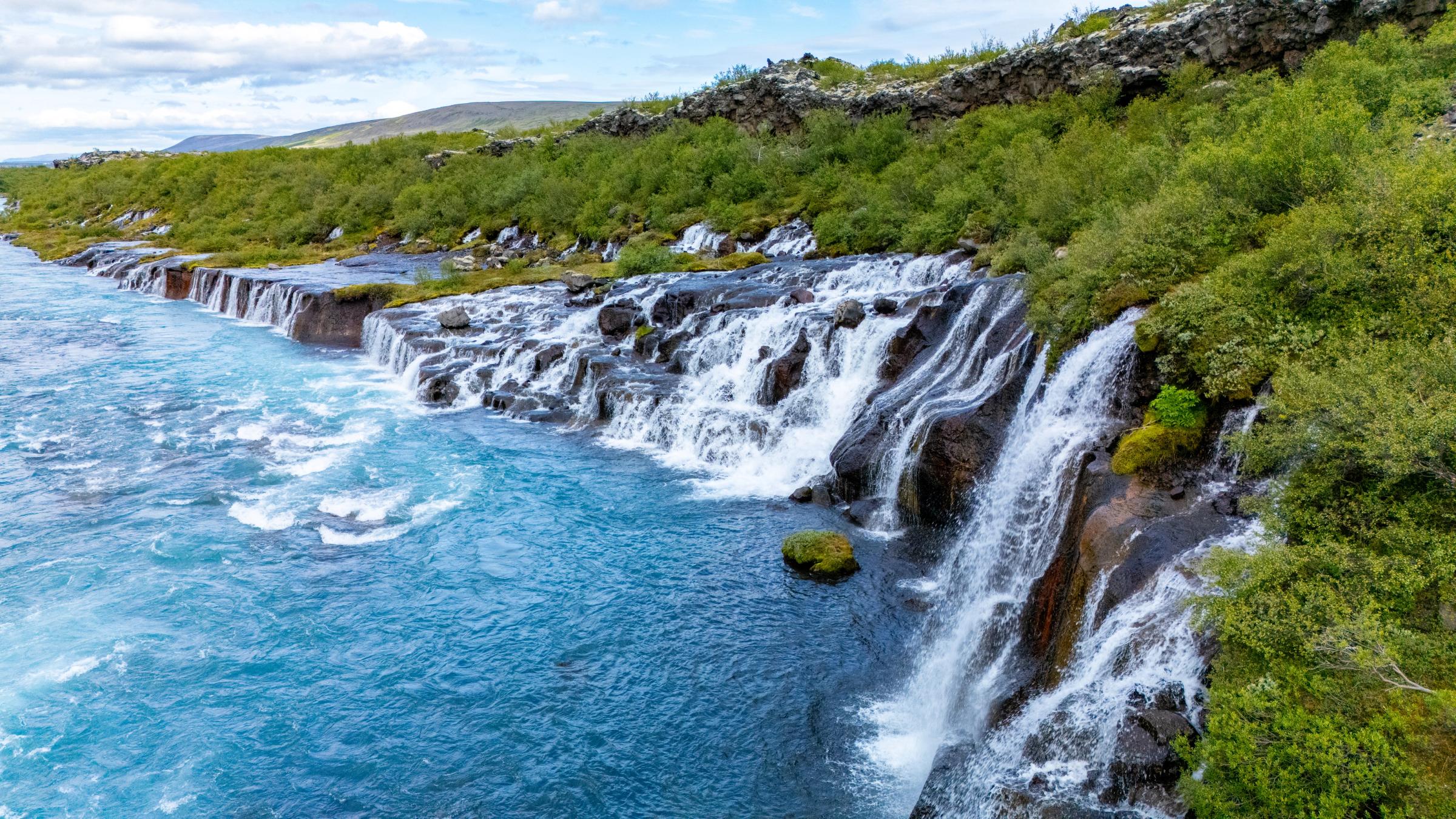 Hraunfossar waterfalls in Iceland, with a turquoise river flowing over lava formations surrounded by green foliage.