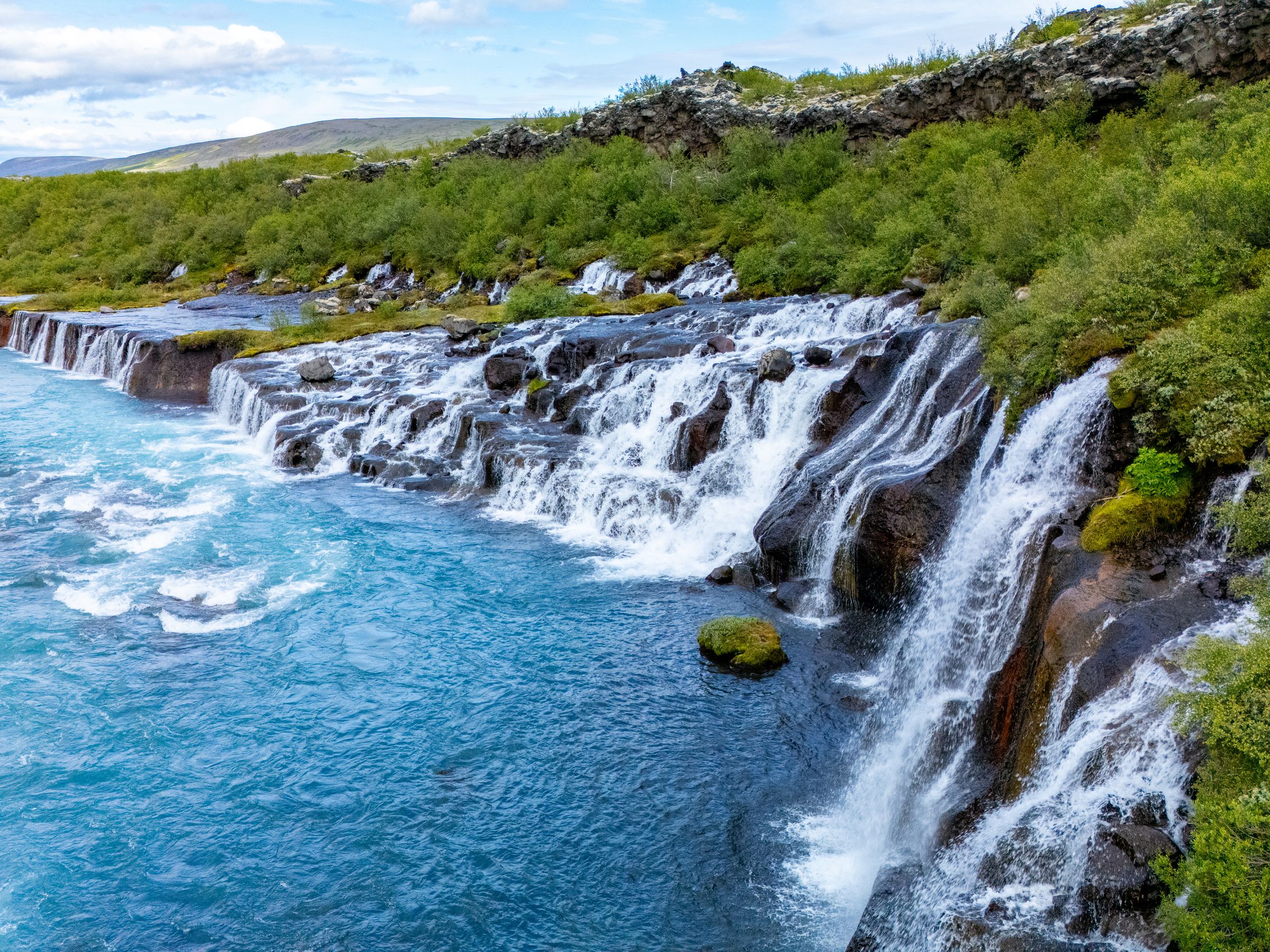 Hraunfossar waterfalls in Iceland, with a turquoise river flowing over lava formations surrounded by green foliage.