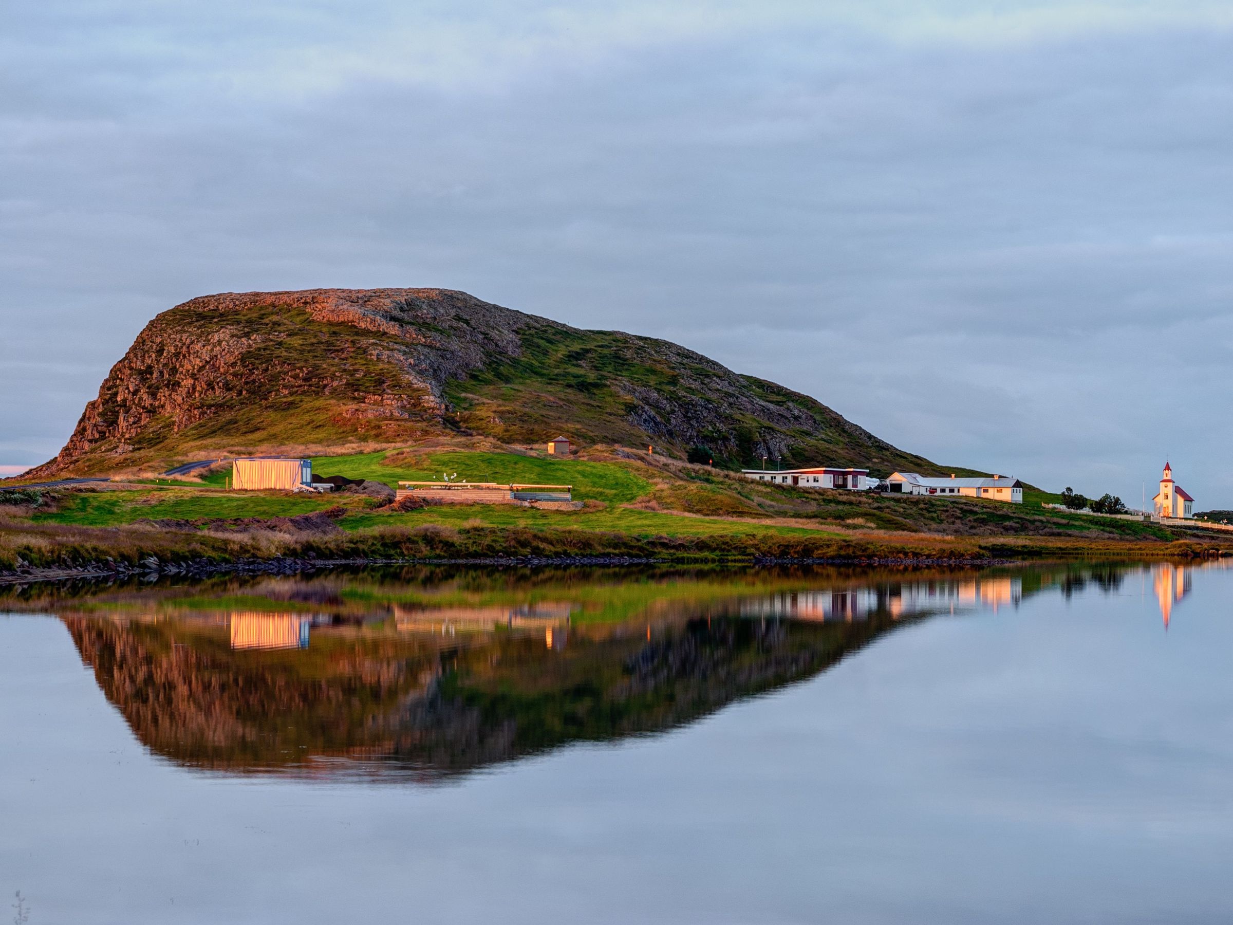 A grassy, rocky hill with a small settlement and a red-roofed church, all perfectly reflected in still water at sunset.