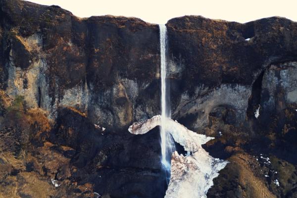 A waterfall cascades down a dark, rocky cliff face with snow and ice at its base.