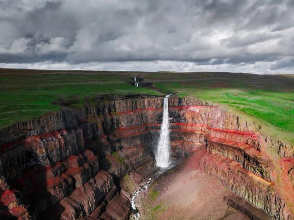 A tall waterfall cascades over dramatic red-layered cliffs into a canyon, with a green plateau above under a cloudy sky.