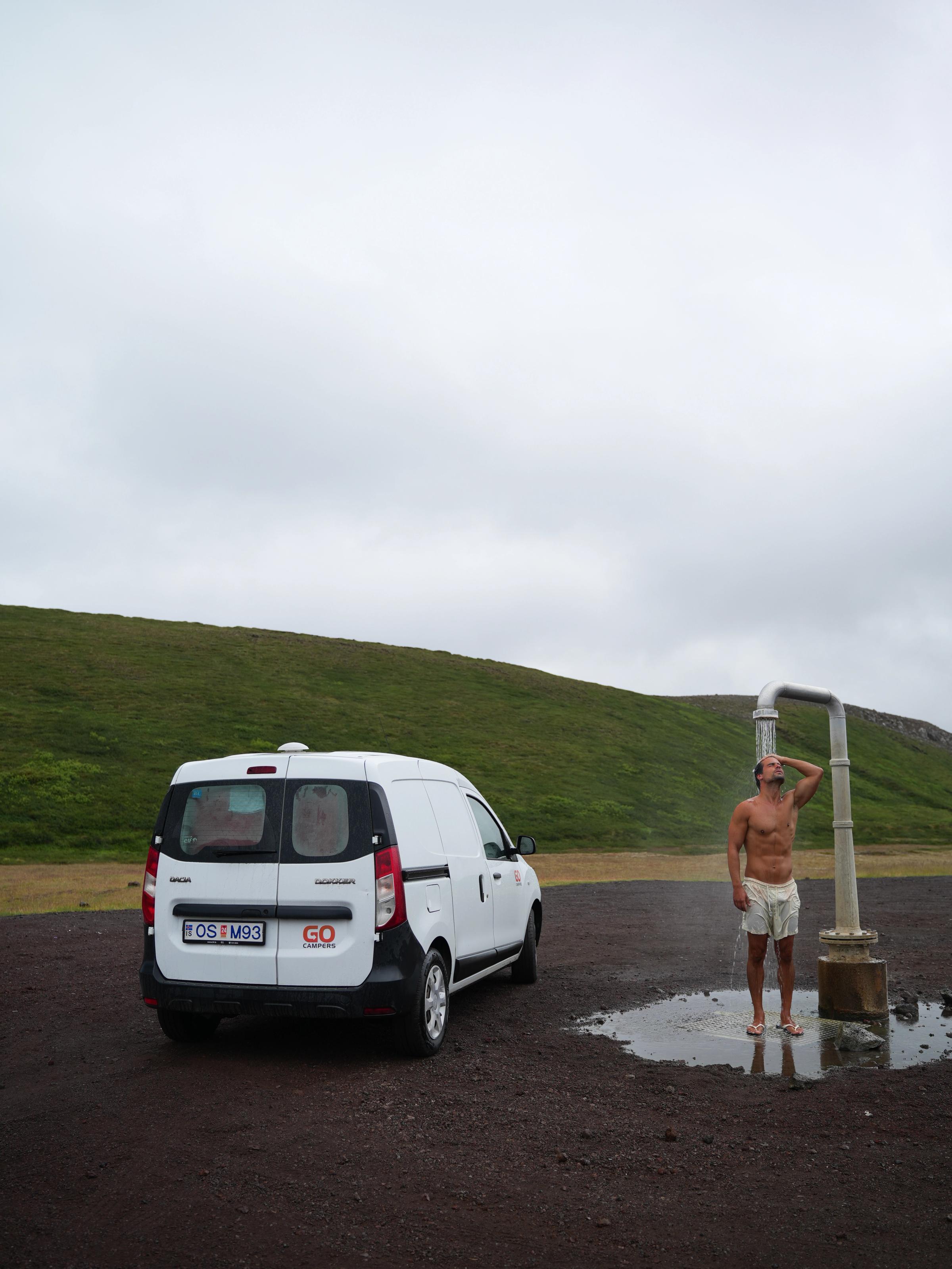 Ein kompakter, günstiger Camper steht auf einem Schotterplatz neben einer Außendusche, die gerade benutzt wird, Campervan-Vermietung Island
