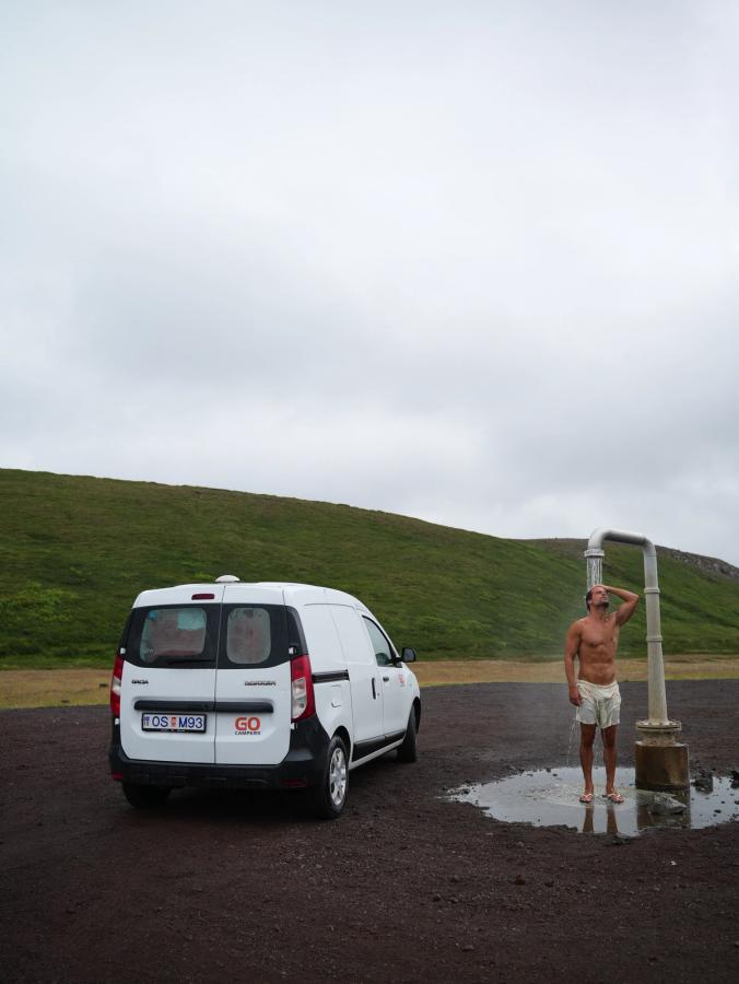 Ein kompakter, günstiger Camper steht auf einem Schotterplatz neben einer Außendusche, die gerade benutzt wird, Campervan-Vermietung Island