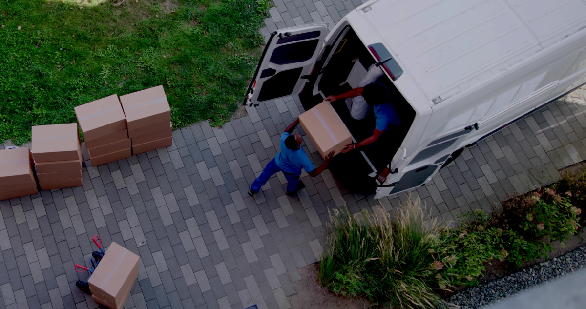 Overhead view of two movers loading a cardboard box into a white delivery van, with more boxes on the ground.