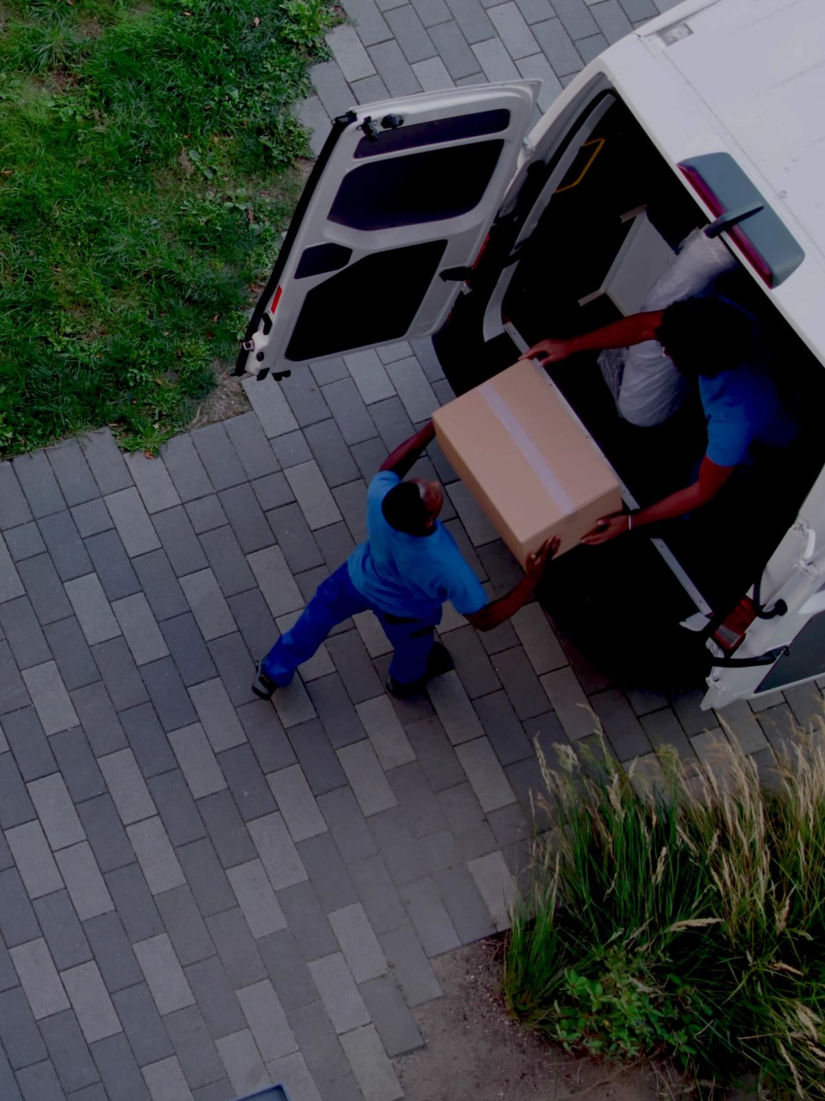 Overhead view of two movers loading a cardboard box into a white delivery van, with more boxes on the ground.