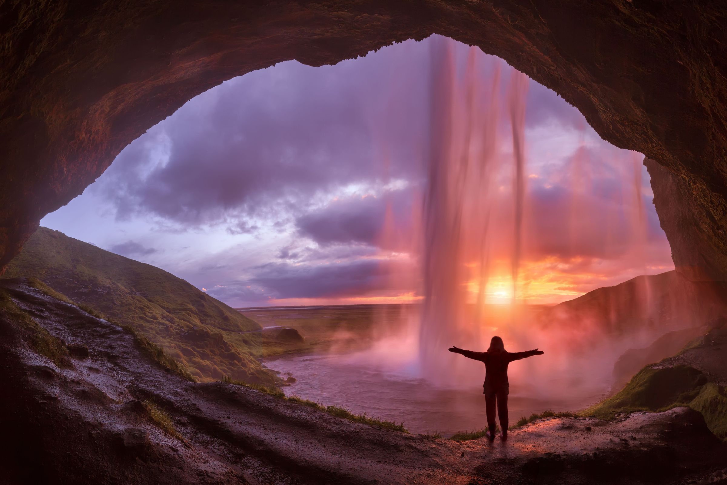 a woman standing behind a waterfall at sunset