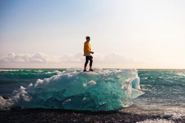 A person in a yellow hoodie stands on a blue ice chunk on a black sand beach by the ocean.