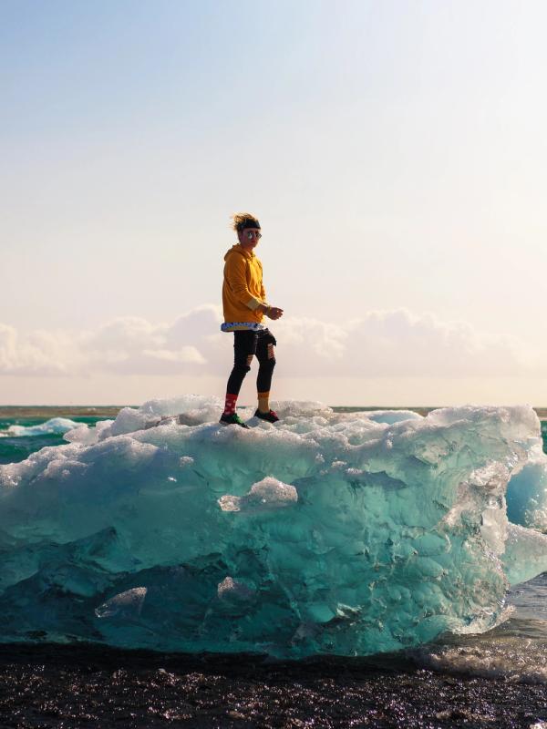 A person in a yellow jacket stands on a large blue ice chunk on a black sand beach with the ocean behind them.