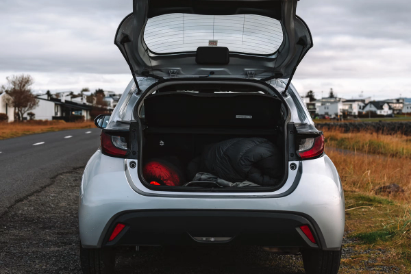 A silver hatchback car with its trunk open, revealing a red bag and dark roll inside, parked on a gravel road under a cloudy sky.