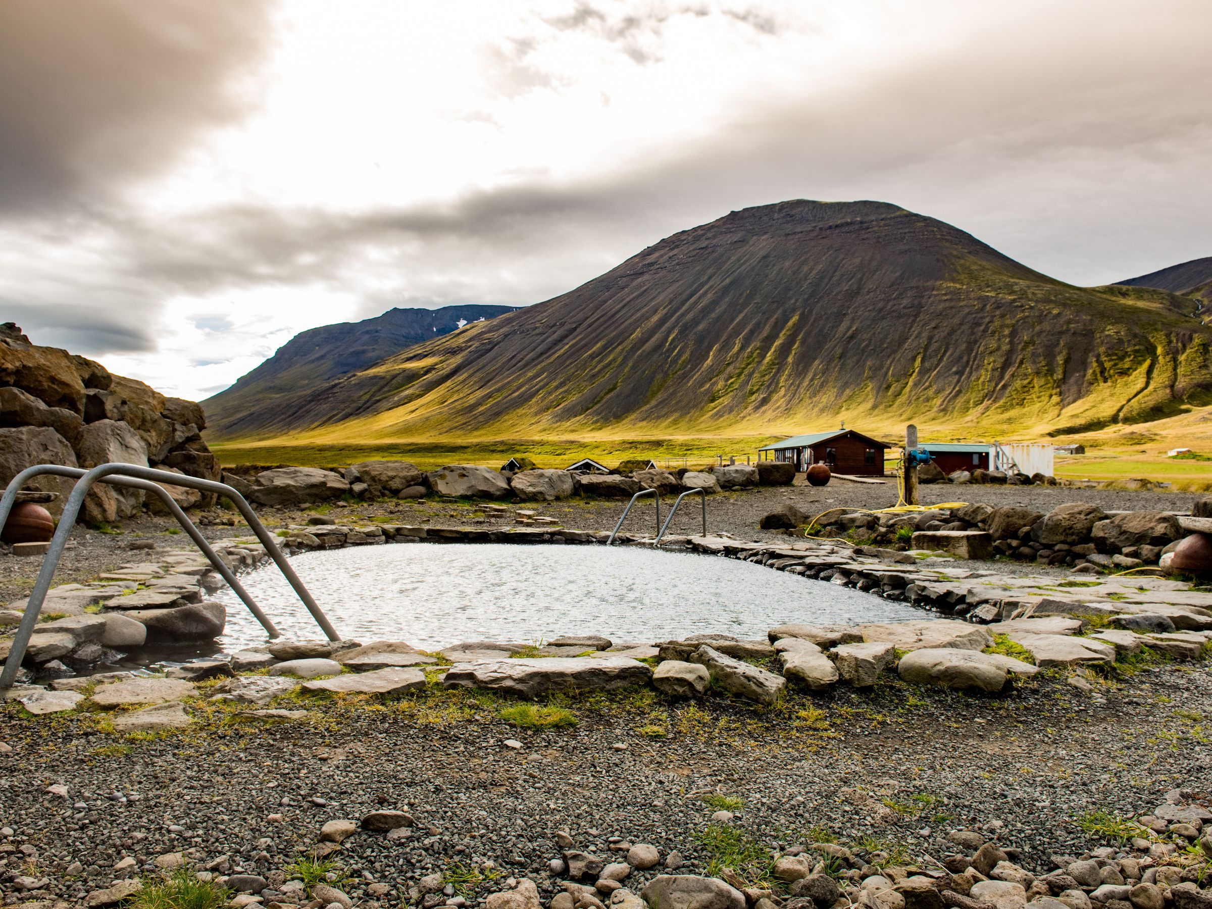 Stone-walled geothermal hot spring at Grettislaug in North Iceland, with mountain views under a cloudy sky