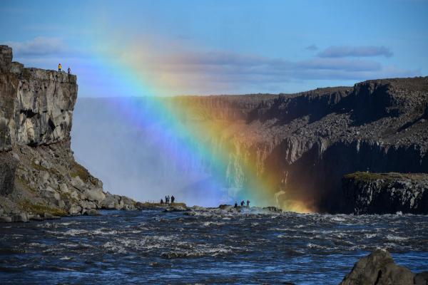 A vibrant rainbow arcs over a powerful waterfall, with spectators on rocky cliffs.