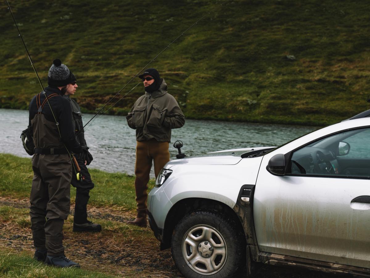 Escena de pesca con un Dacia Duster Dacia Duster 4x4 de alquiler en Islandia aparcado junto a un río, con dos personas sosteniendo cañas de pescar