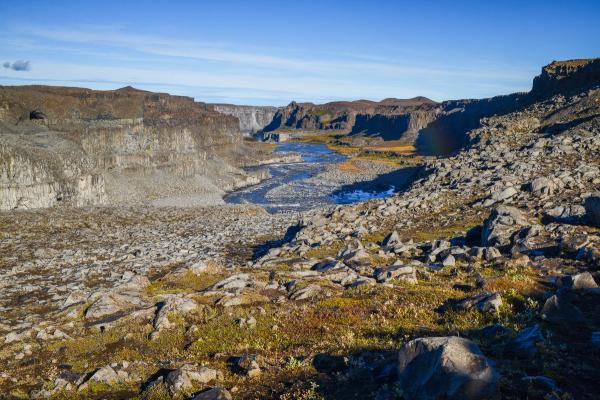 A wide, rugged canyon with a rocky river, high cliffs, sparse vegetation, and a faint rainbow under a clear blue sky.