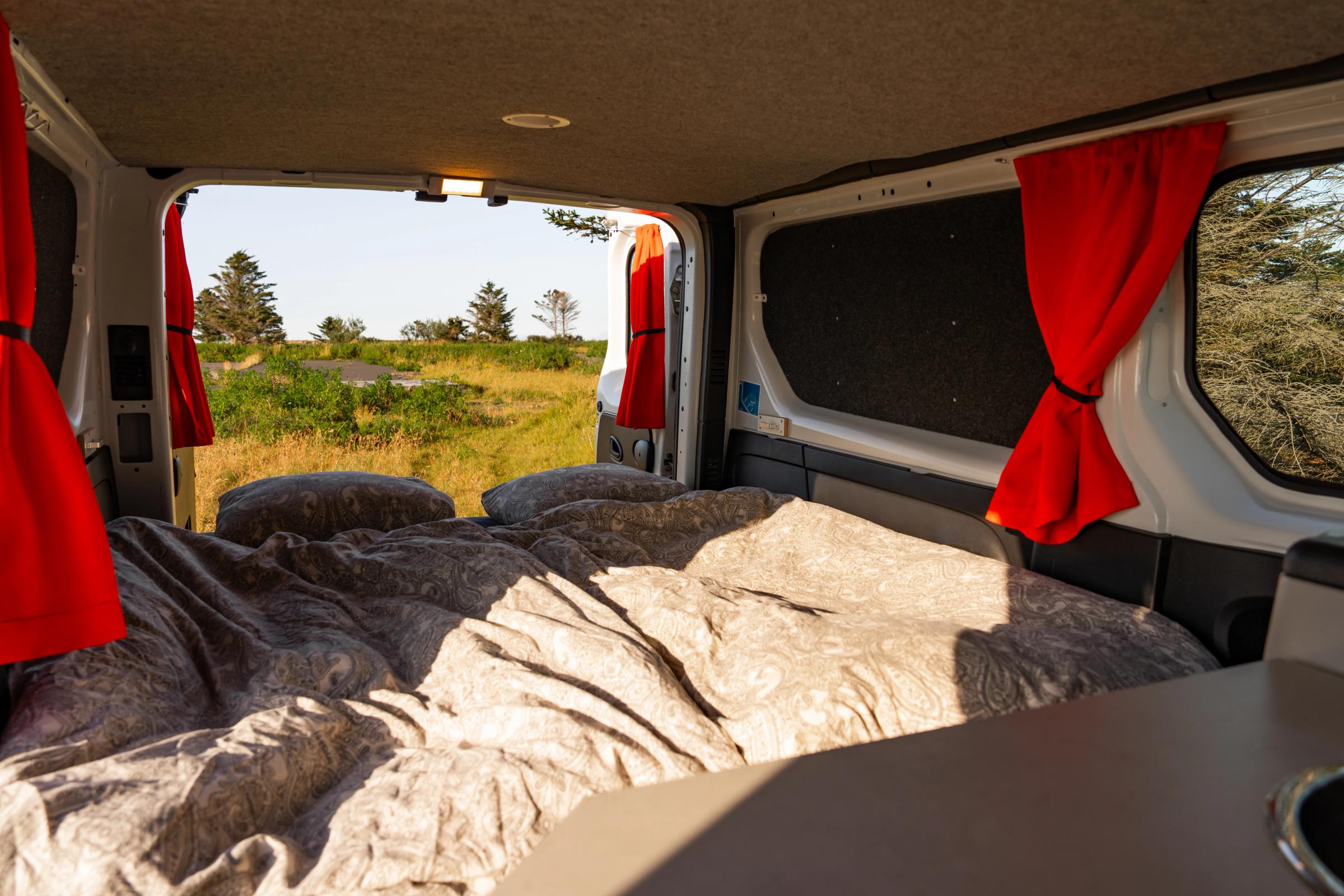 Bed made up with duvet and pillows, with rear doors open to a grassy landscape