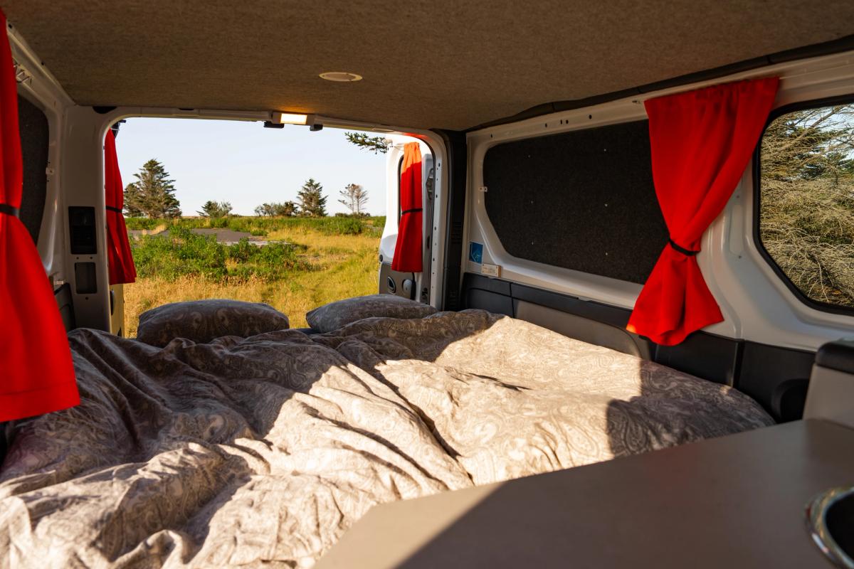 Bed made up with duvet and pillows, with rear doors open to a grassy landscape