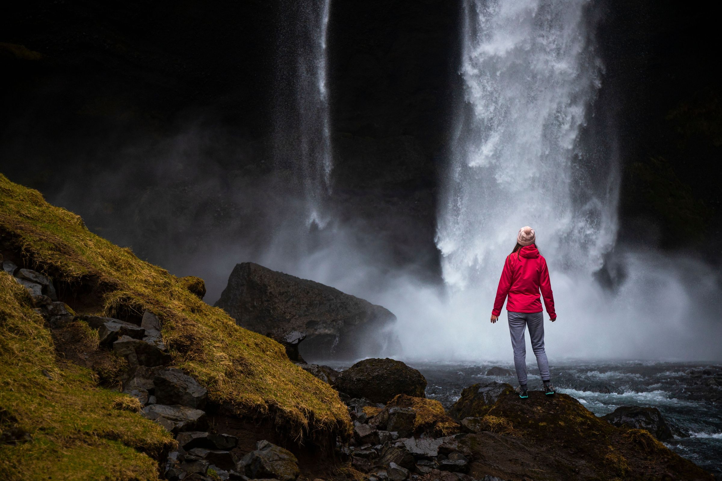 a woman in a red jacket is standing in front of a waterfall .